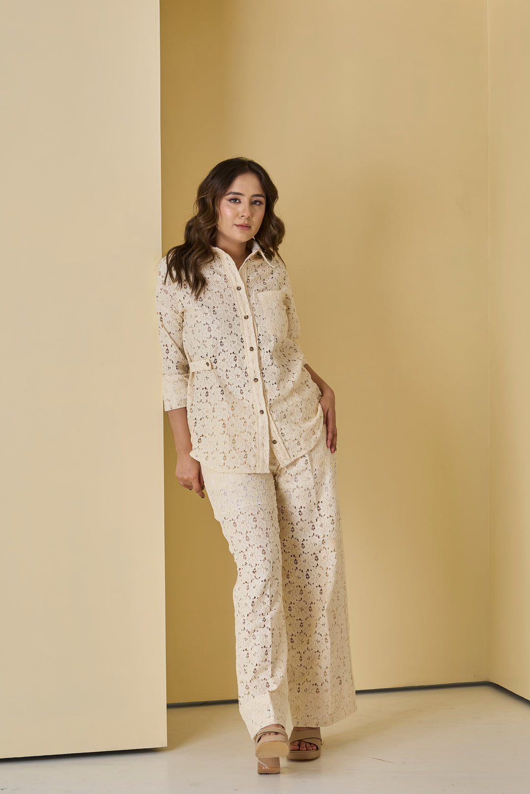 Woman wearing a beige lace outfit standing against a beige wall.