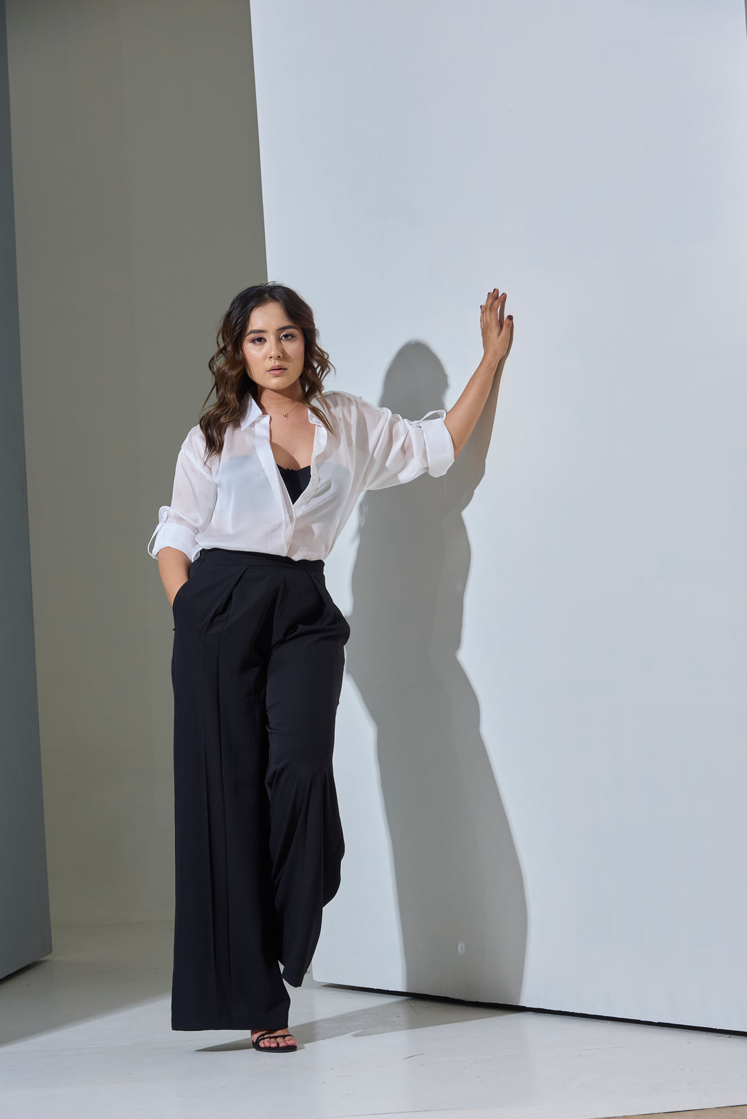 Woman in a white blouse and black pants standing against a plain wall with a soft shadow.