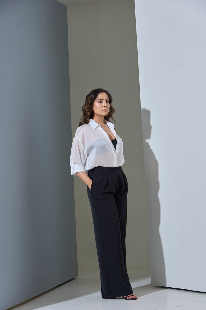 Woman in a white blouse and black pants standing against a plain wall.