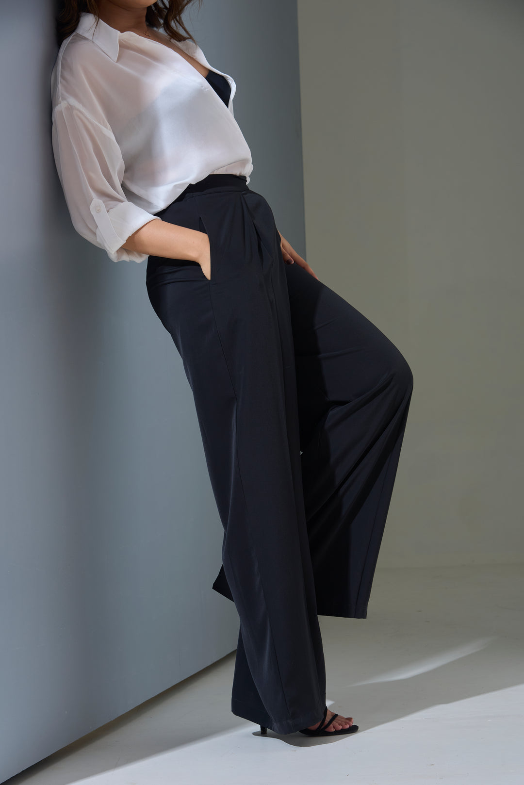 Woman in a white blouse and black pants standing against a plain wall with a soft shadow.
