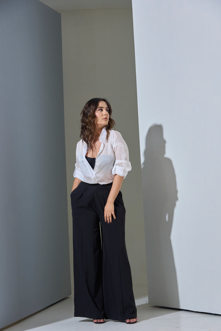 Woman in a white blouse and black pants standing against a plain wall with a soft shadow.
