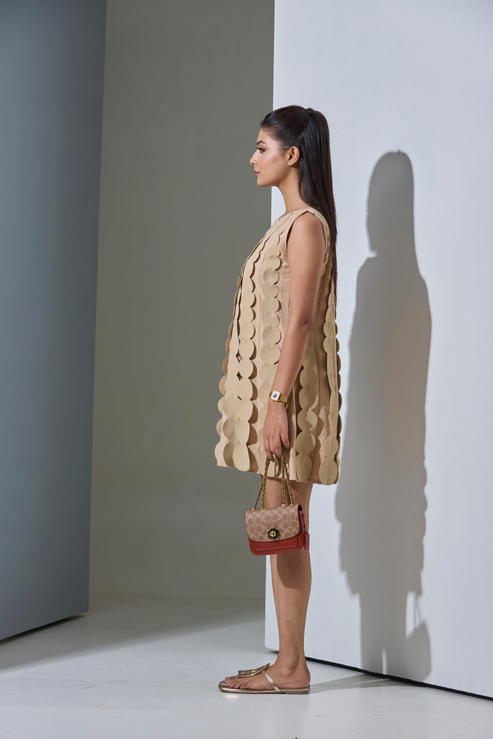 Woman wearing a beige dress with a textured pattern, standing against a white wall.