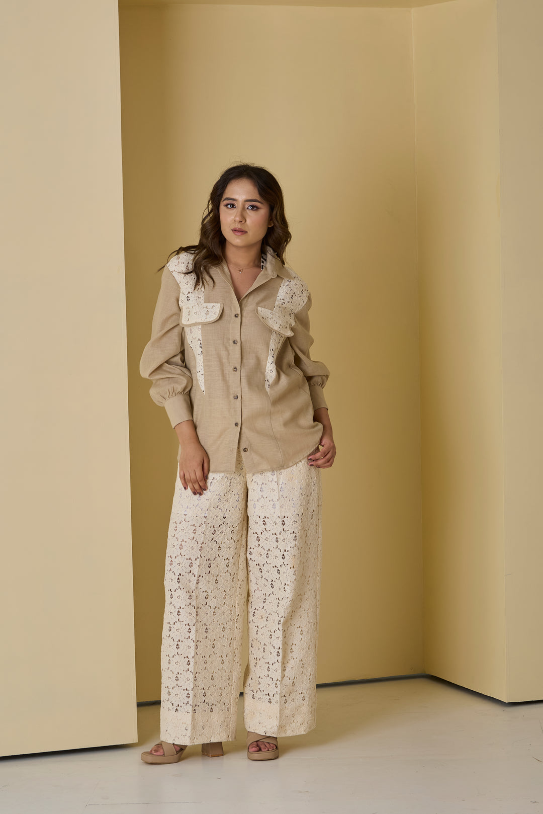 Woman wearing a beige shirt with lace details and white patterned pants against a beige wall.